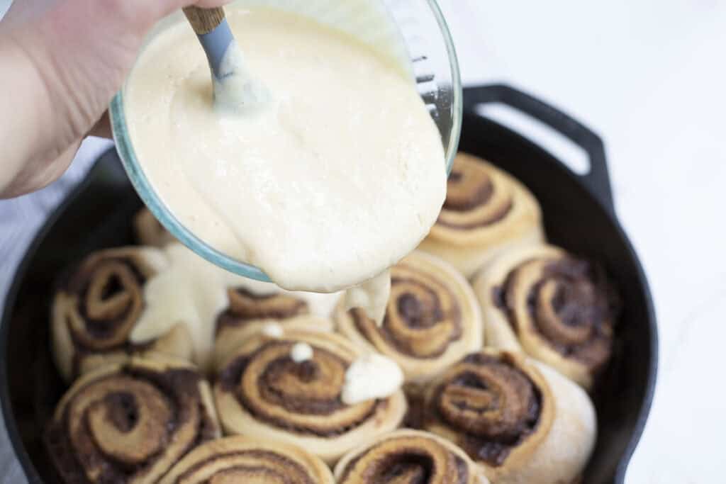 pouring cream cheese topping over cinnamon rolls in a cast iron skillet