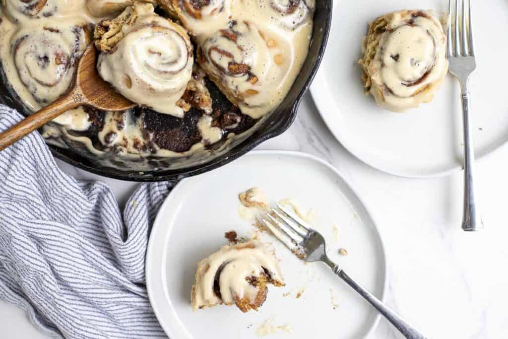 two plates with sourdough discard cinnamon rolls topped with cream cheese topping on a blue and white stripped towel. A cast iron skillet with more rolls is in the background