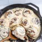 cast iron skillet with sourdough cinnamon rolls topped with cream cheese topping. A wooden spoon is in the skillet with a cinnamon roll on top.