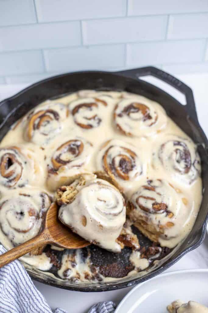 cast iron skillet with sourdough cinnamon rolls topped with cream cheese topping. A wooden spoon is in the skillet with a cinnamon roll on top.