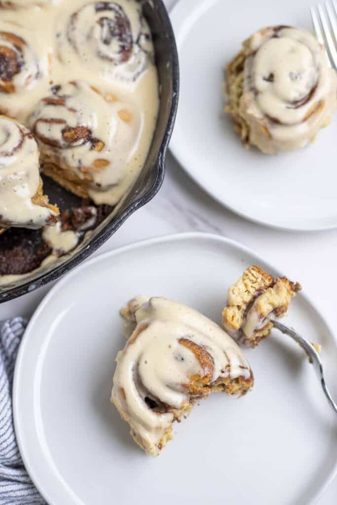 overhead photo of a sourdough cinnamon roll with a portion taken out and on a fork on the plate. Another roll is on another plate and a skillet with more rolls are in the background