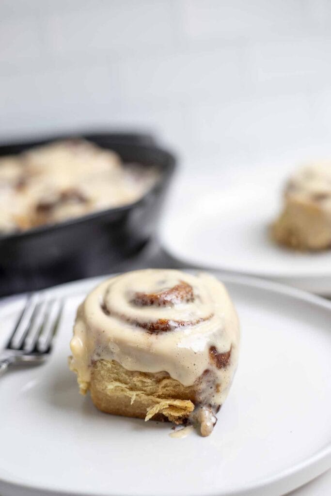 a sourdough cinnamon roll on a cream colored plate with a fork. Another roll on a plate and cast iron skillet with more rolls is in the background