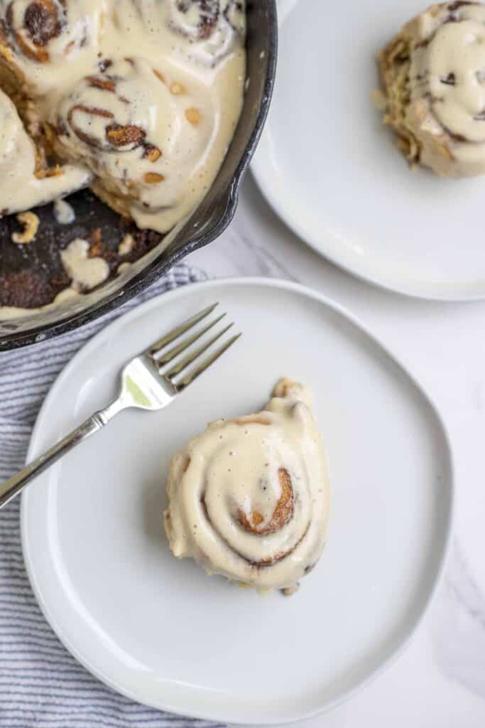 overhead photo of sourdough discard cinnamon roll on a cream plate with a fork. Another plate with a roll and a cast iron skillet with more rolls is in the background