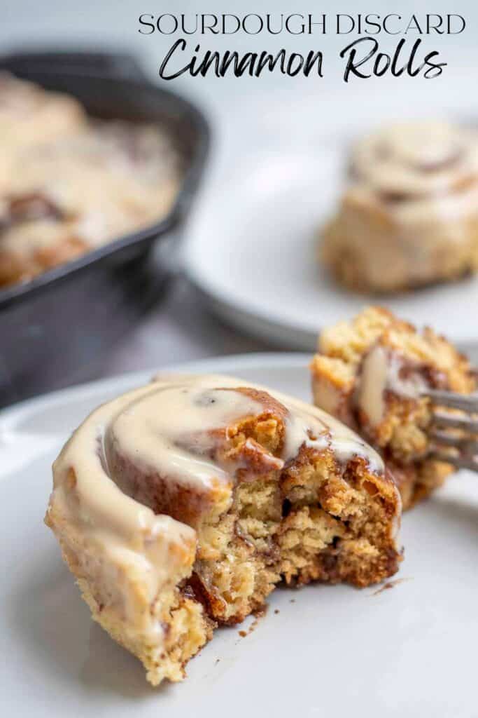 Sourdough discard cinnamon roll cut in half on a plate. Another cinnamon roll on a plate and a cast iron skillet with more cinnamon rolls are in the background