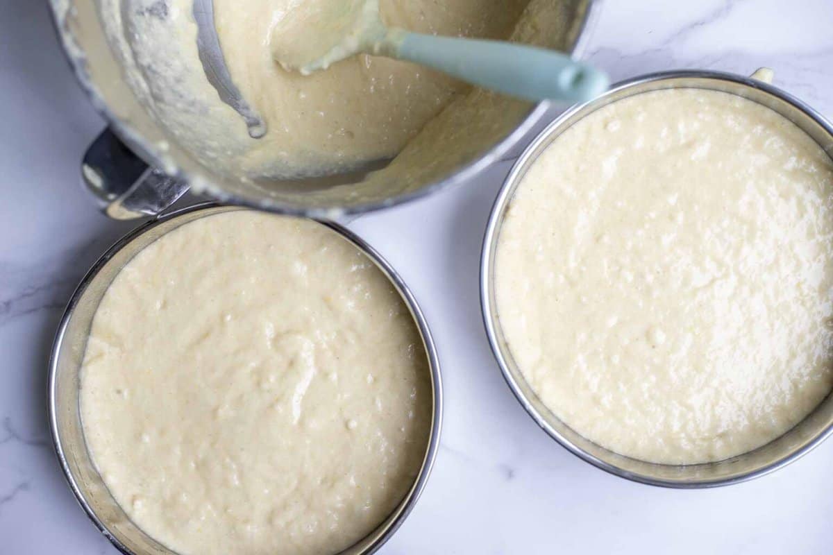 Lemon sourdough cake batter poured into two round cake pans next to the bowl of a stand mixer with remaining batter on a white countertop.