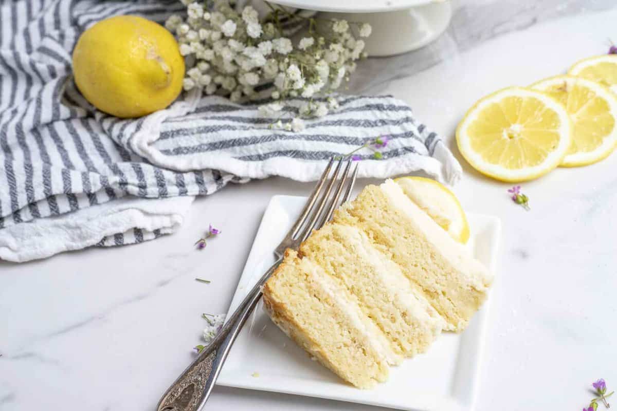 A slice of layered lemon sourdough cake on a white plate with a silver fork surrounded by lemon slices on a white countertop.