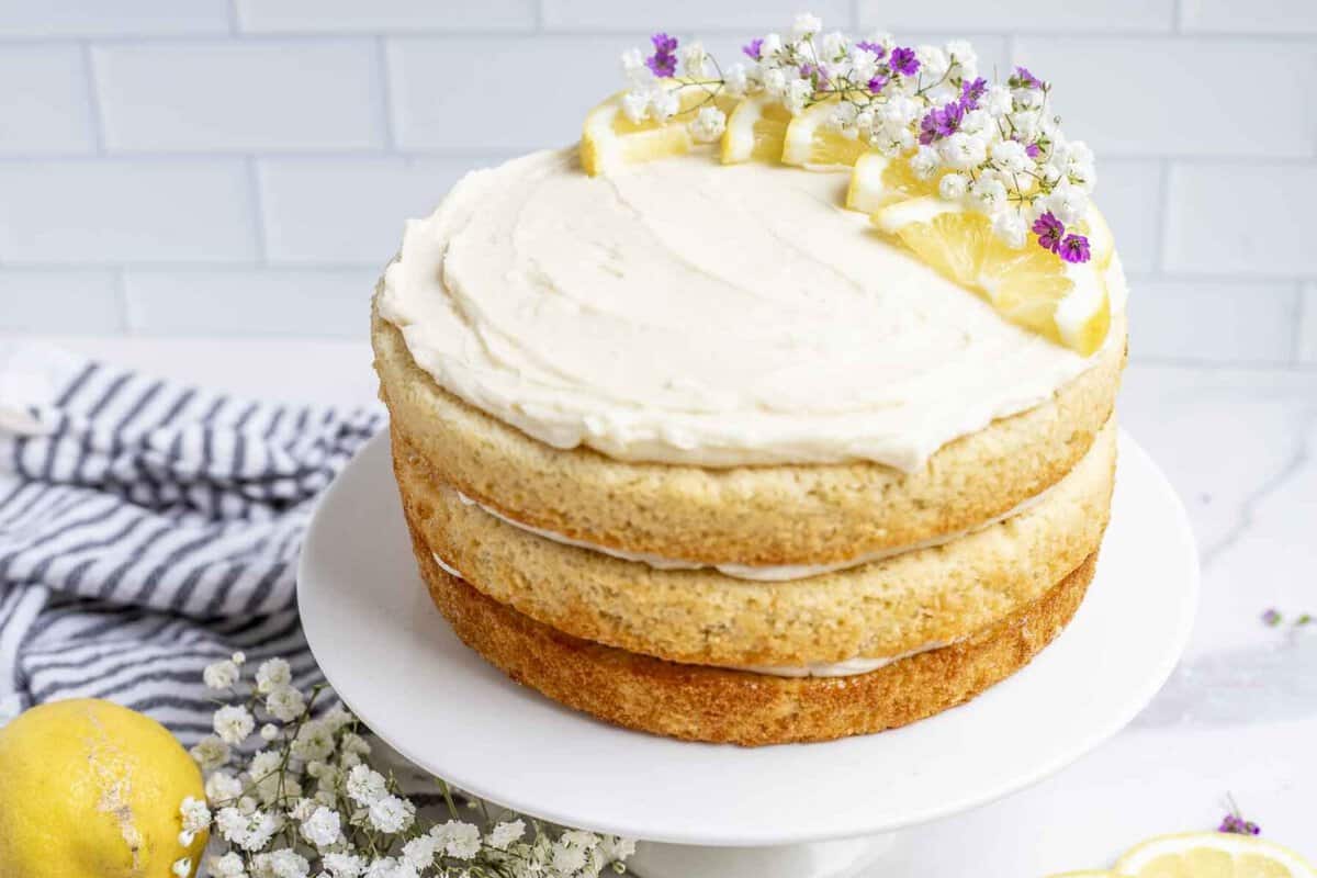 Layered sourdough lemon cake with white icing and lemon decorations on a white cake plate with a blue checkered towel and full lemon on the side