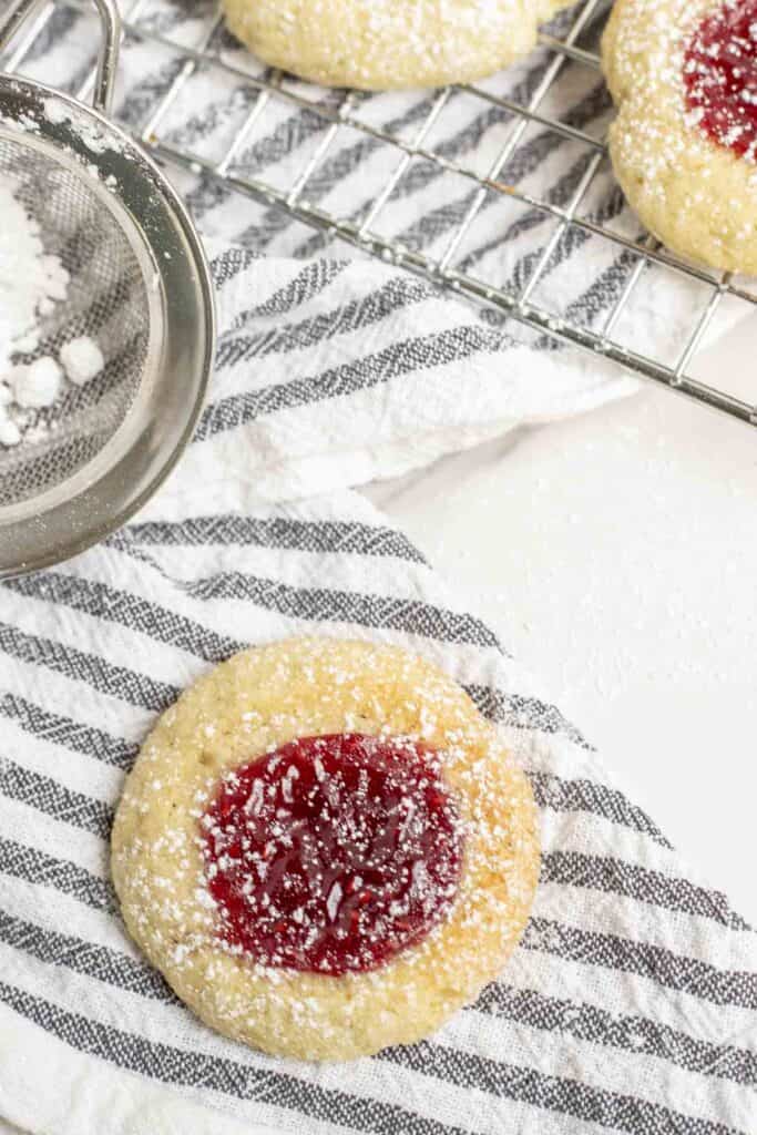 one sugar cookie with jam in the center on a white and black stripped towel with more cookies on a wire rack in the background