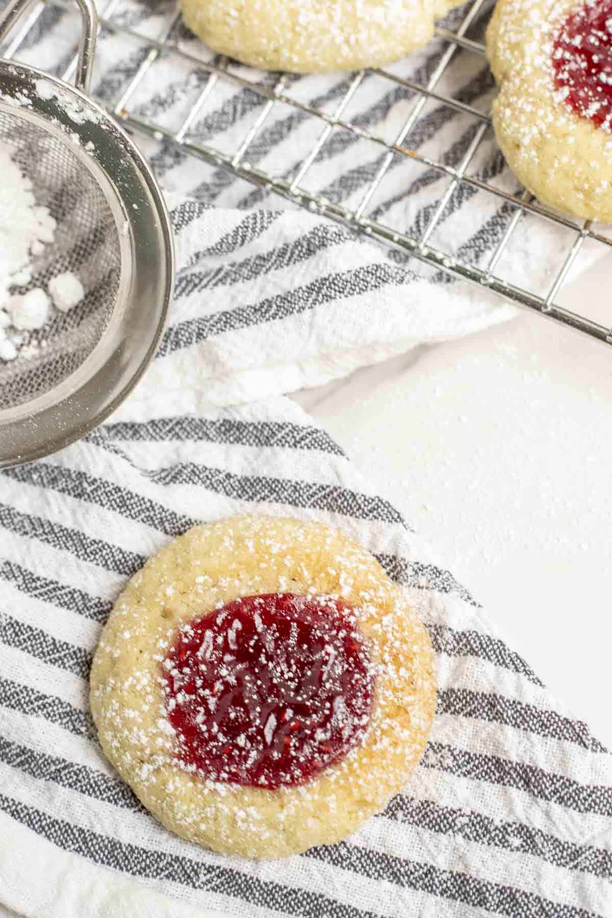 one sugar cookie with jam in the center on a white and black stripped towel with more cookies on a wire rack in the background