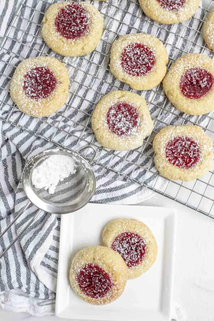 two thumbprint sourdough cookies on a white plate. More cooking on a cooling wrack on top of a white and black stripped towel in the background.