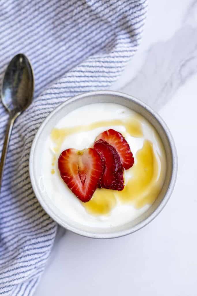 Raw milk yogurt topped with maple syrup and sliced strawberries in a bowl on a white and blue striped towel