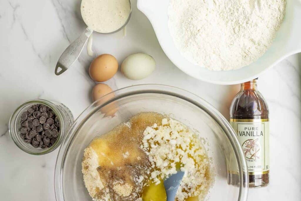 one white bowl with flour, baking soda, baking powder and salt. a bowl of sugar and melted butter, eggs, vanilla extract, a jar of chocolate chips, and a measuring cup of starter on a marble counter