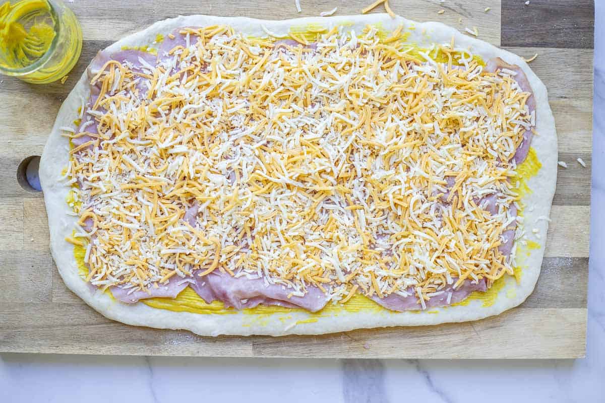 Sourdough ham and cheese roll dough rolled out on a wooden cutting board and brushed with mustard and layered with ham slices and shredded cheese