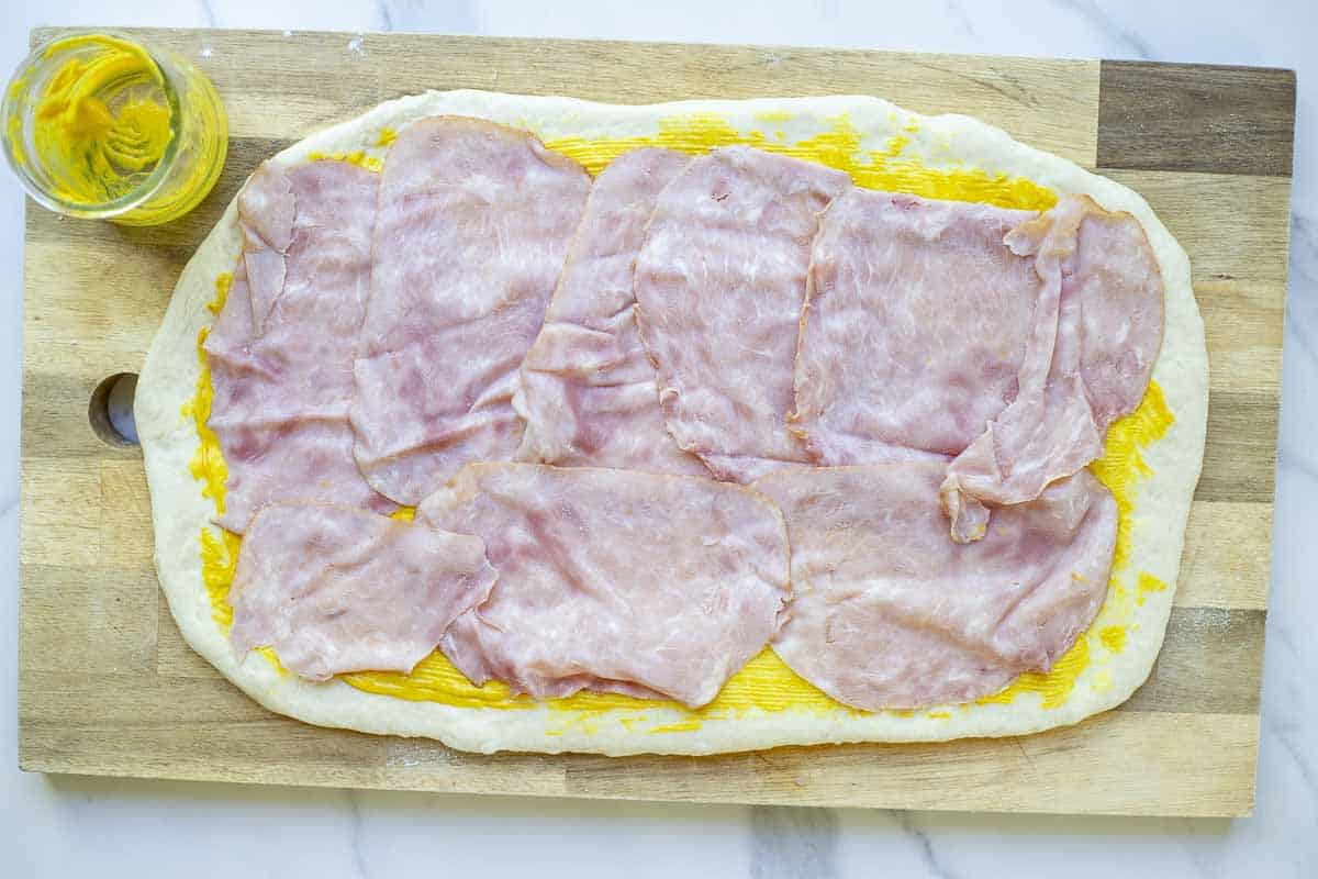 Sourdough ham and cheese roll dough rolled out with mustard brushed on and slices of ham on top on a wooden cutting board