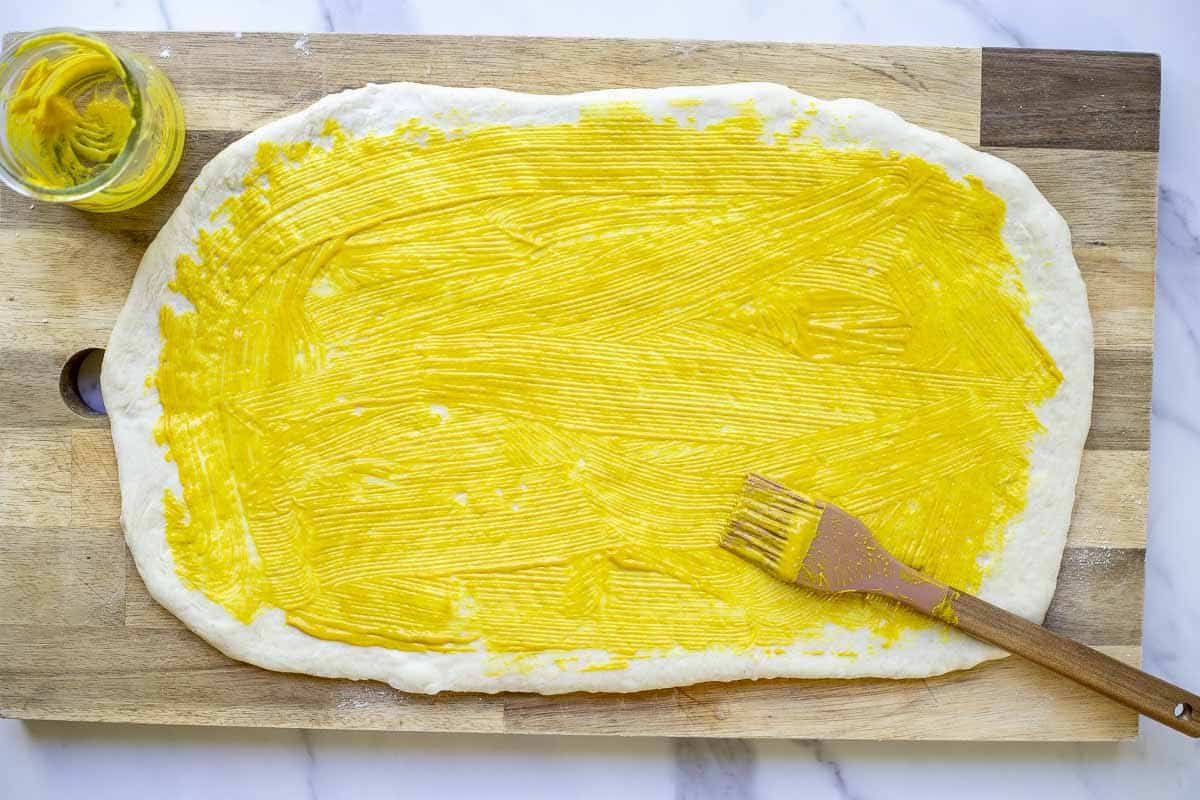 Sourdough ham and cheese roll dough rolled out and brushed with mustard on a wooden cutting board with a pastry brush setting on the side
