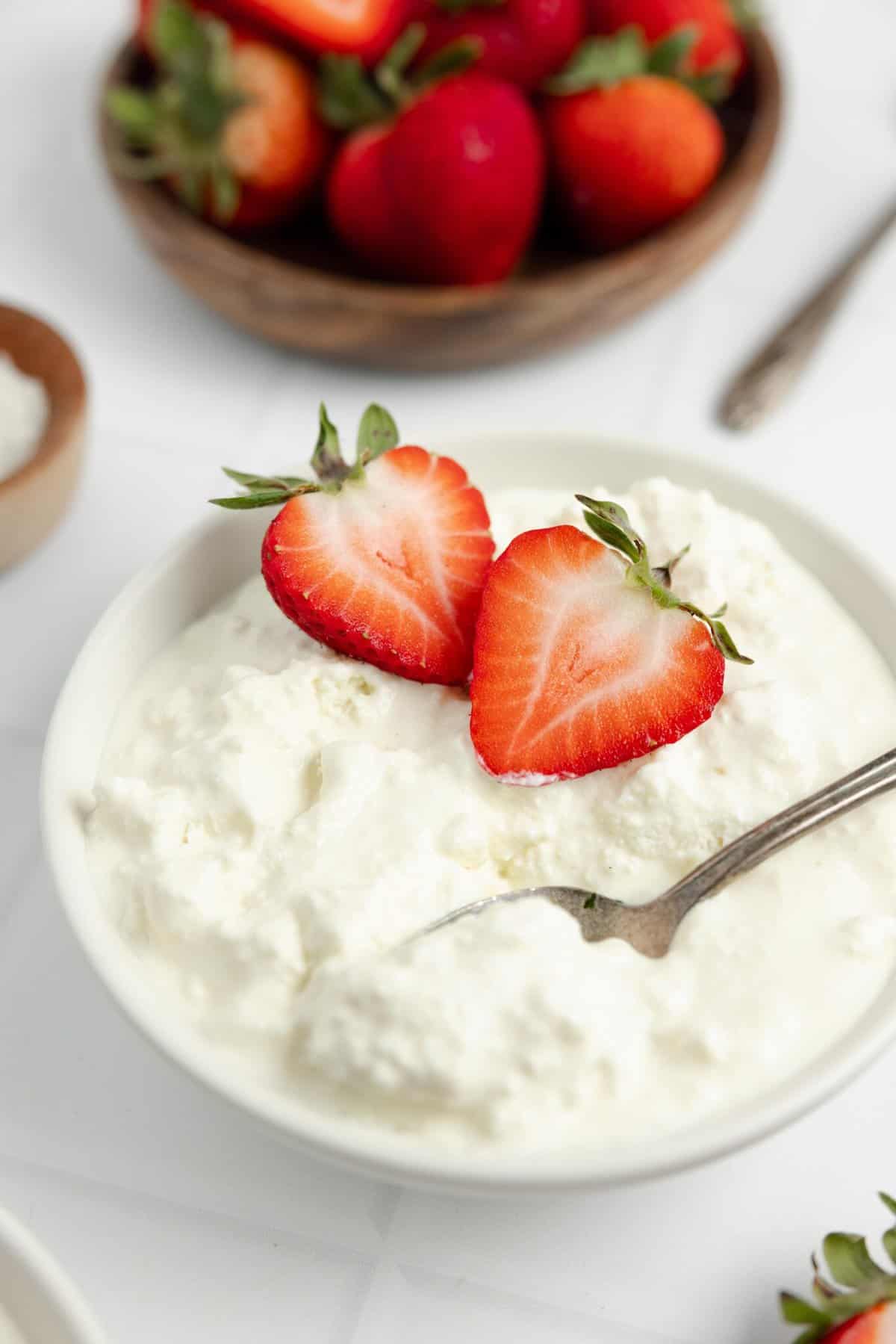 Homemade cottage cheese topped with halved strawberries in a white bowl. More strawberries in a bowl in the background.
