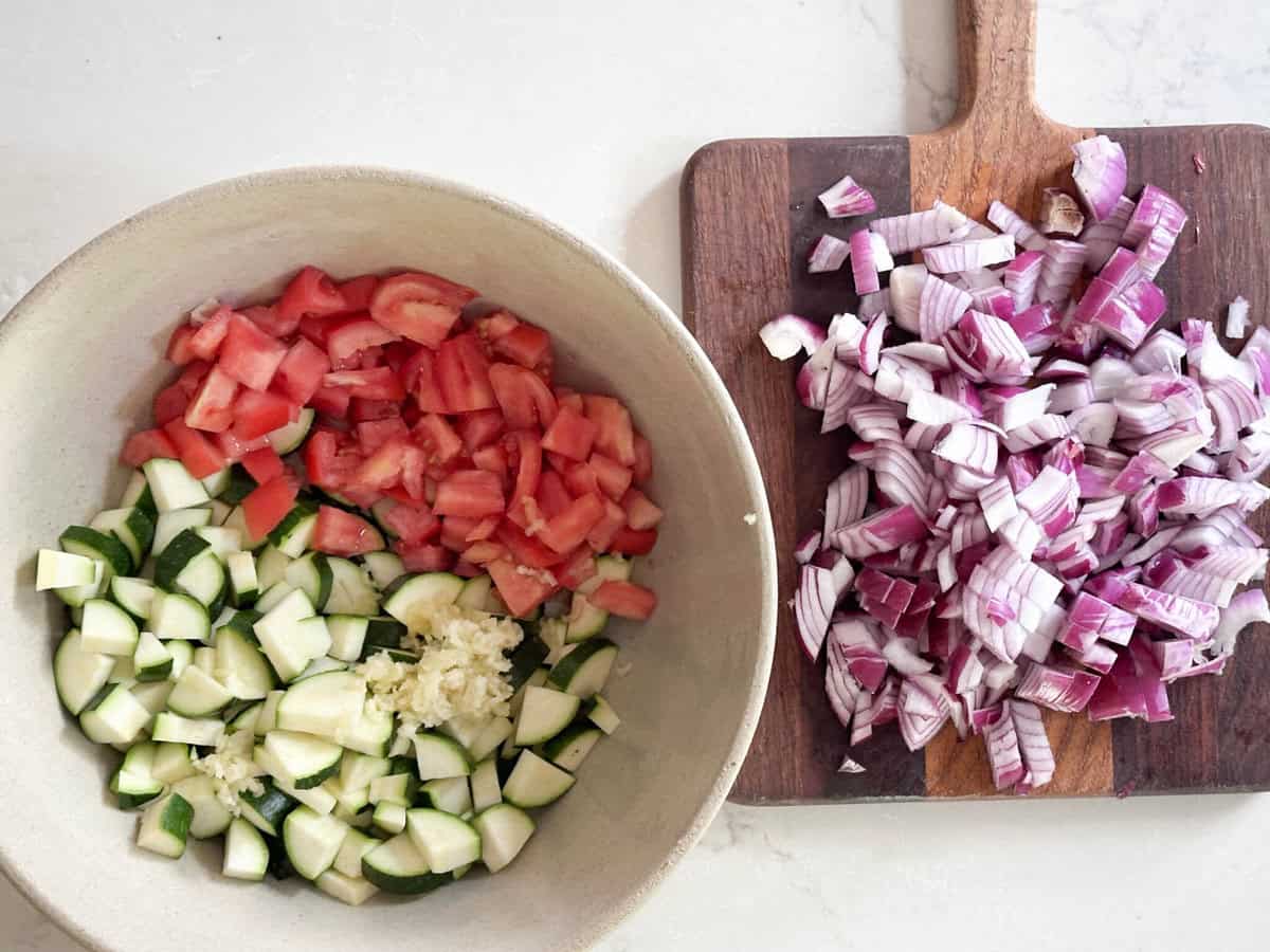 Chopped onion on a cutting board next to a bowl of chopped zucchini and tomato.