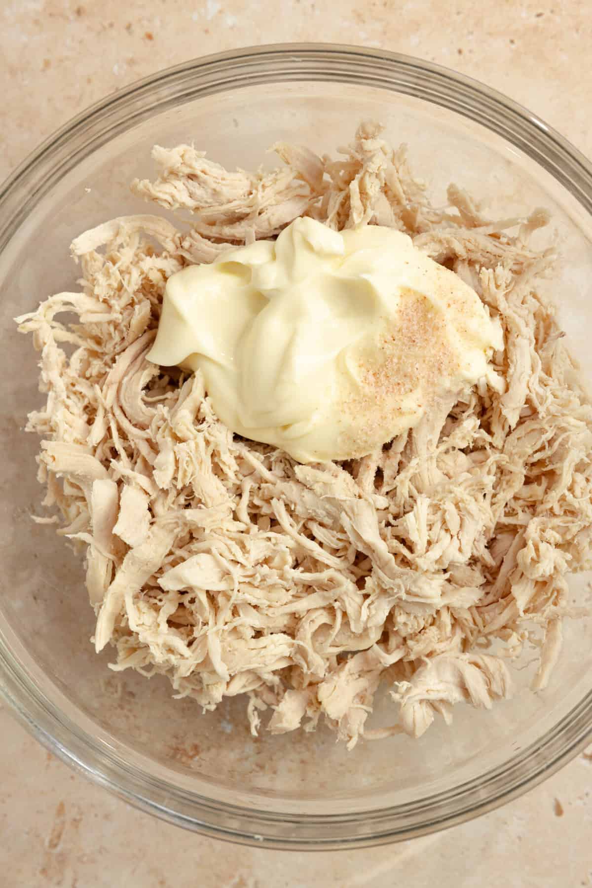 Shredded chicken with a half cup of mayo on top in a clear bowl.