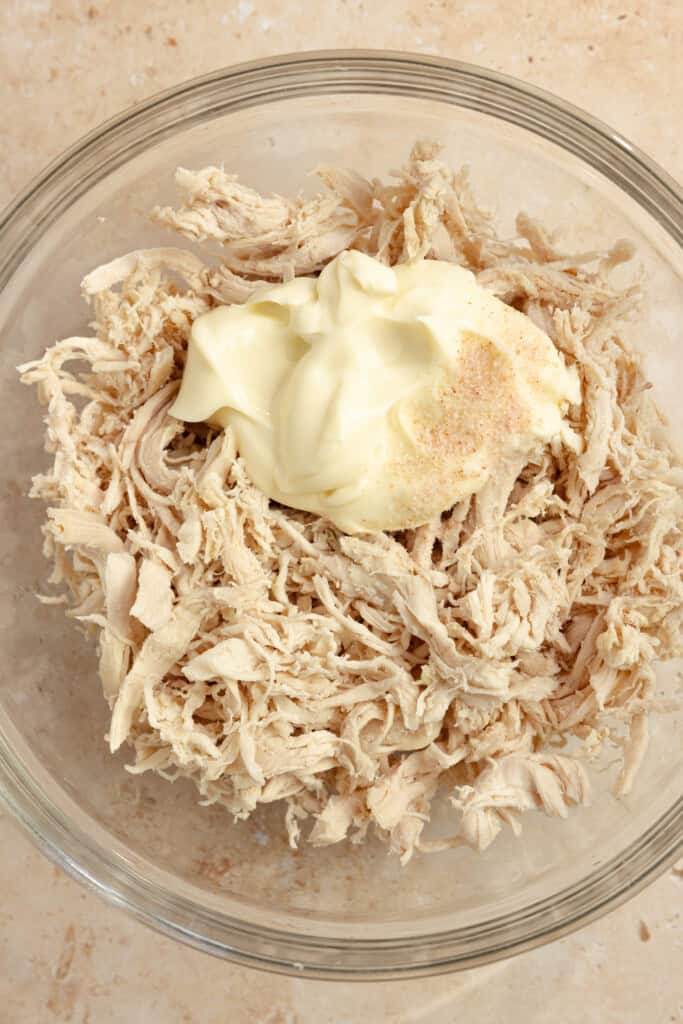 Shredded chicken with a half cup of mayo on top in a clear bowl.