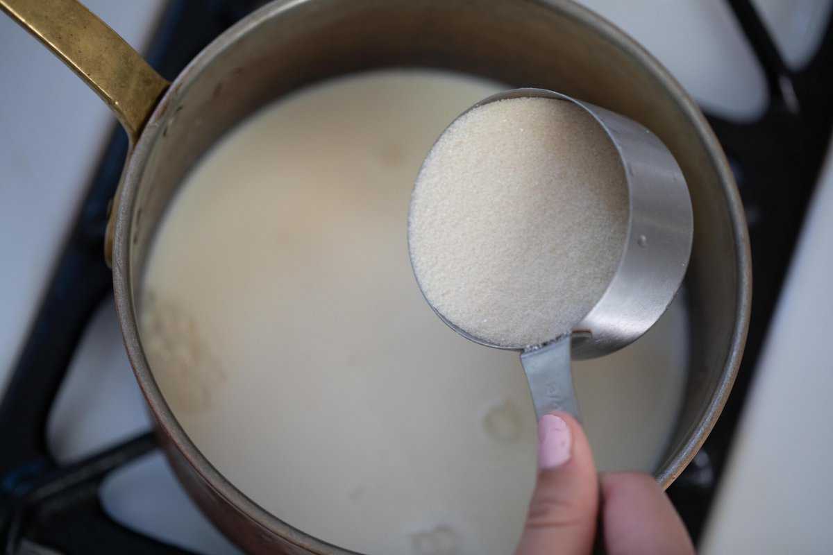 A hand adding a measuring cup full of sugar to a saucepan of milk.