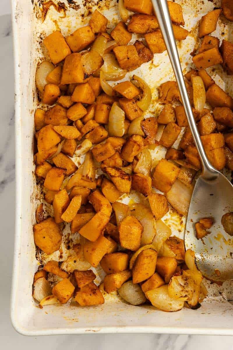 Roasted pumpkin and onion in a white baking dish with a large silver spoon.