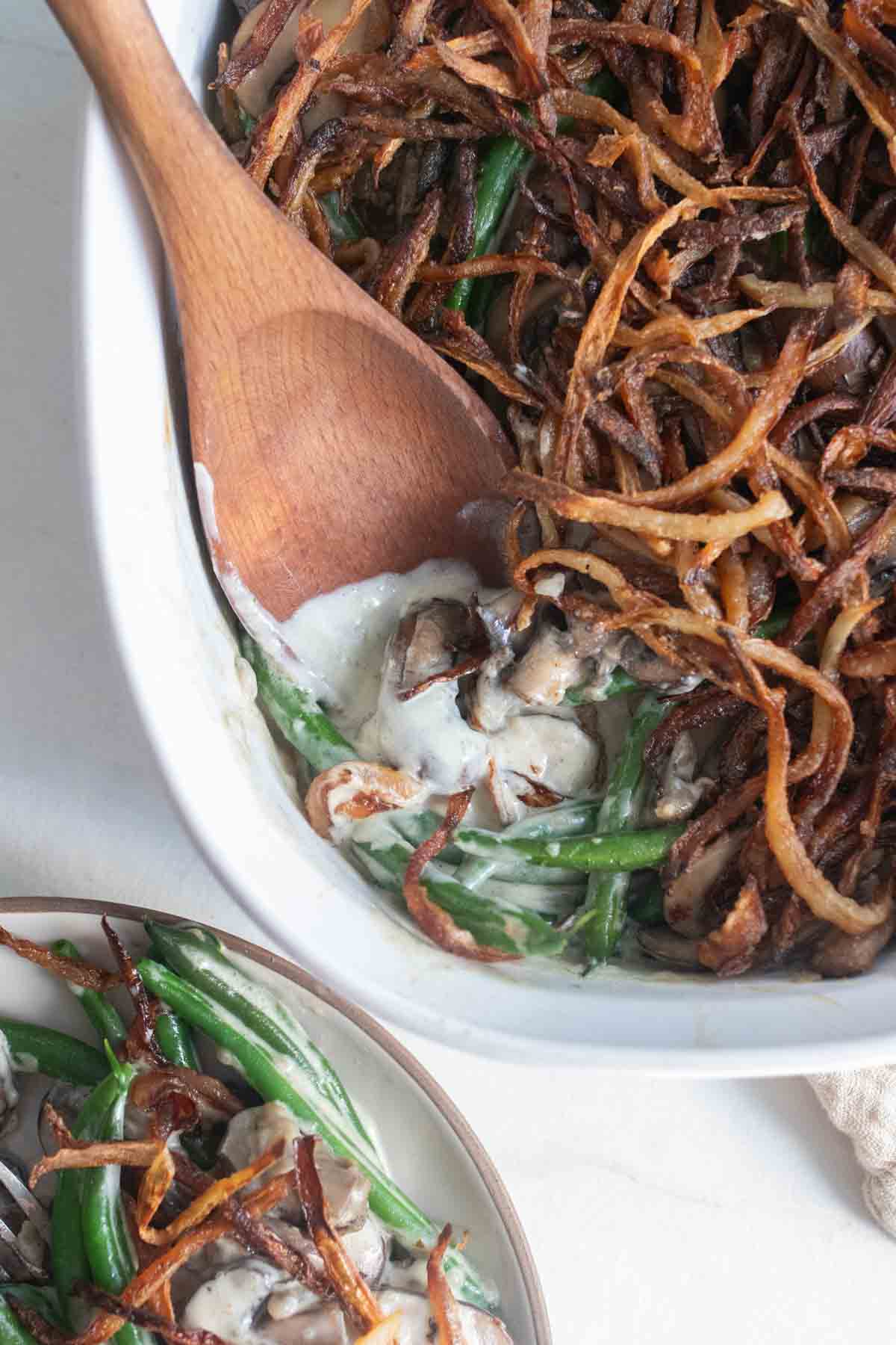 A wooden spoon in a baking dish scooping up one serving of green bean casserole.
