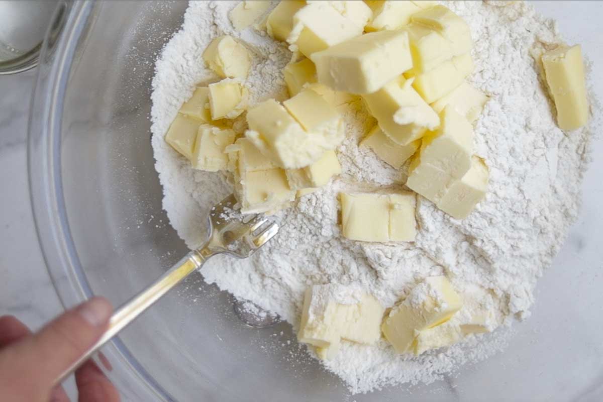 A bowl of flour with cubed butter being added by a fork.