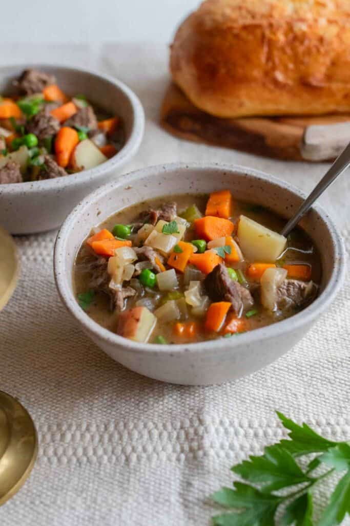 A bowl of beef stew with a loaf of bread in the background.