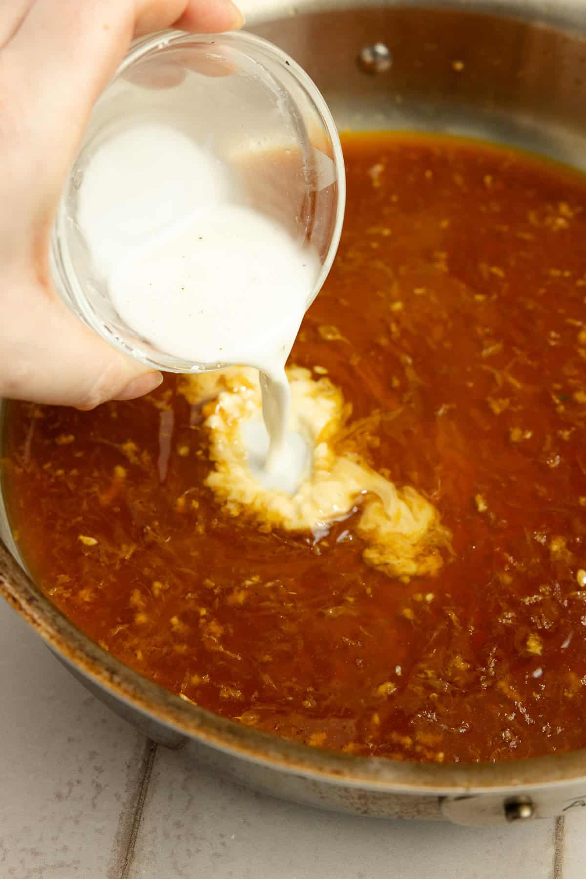 A hand adding a cornstarch slurry to a saucepan of sauce.