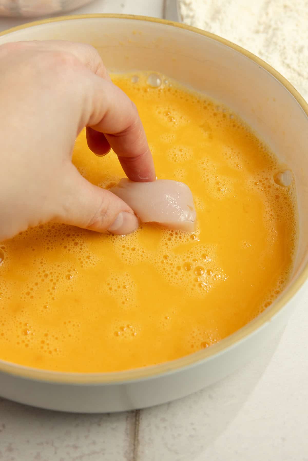 A hand dipping chicken into a bowl of whisked egg.