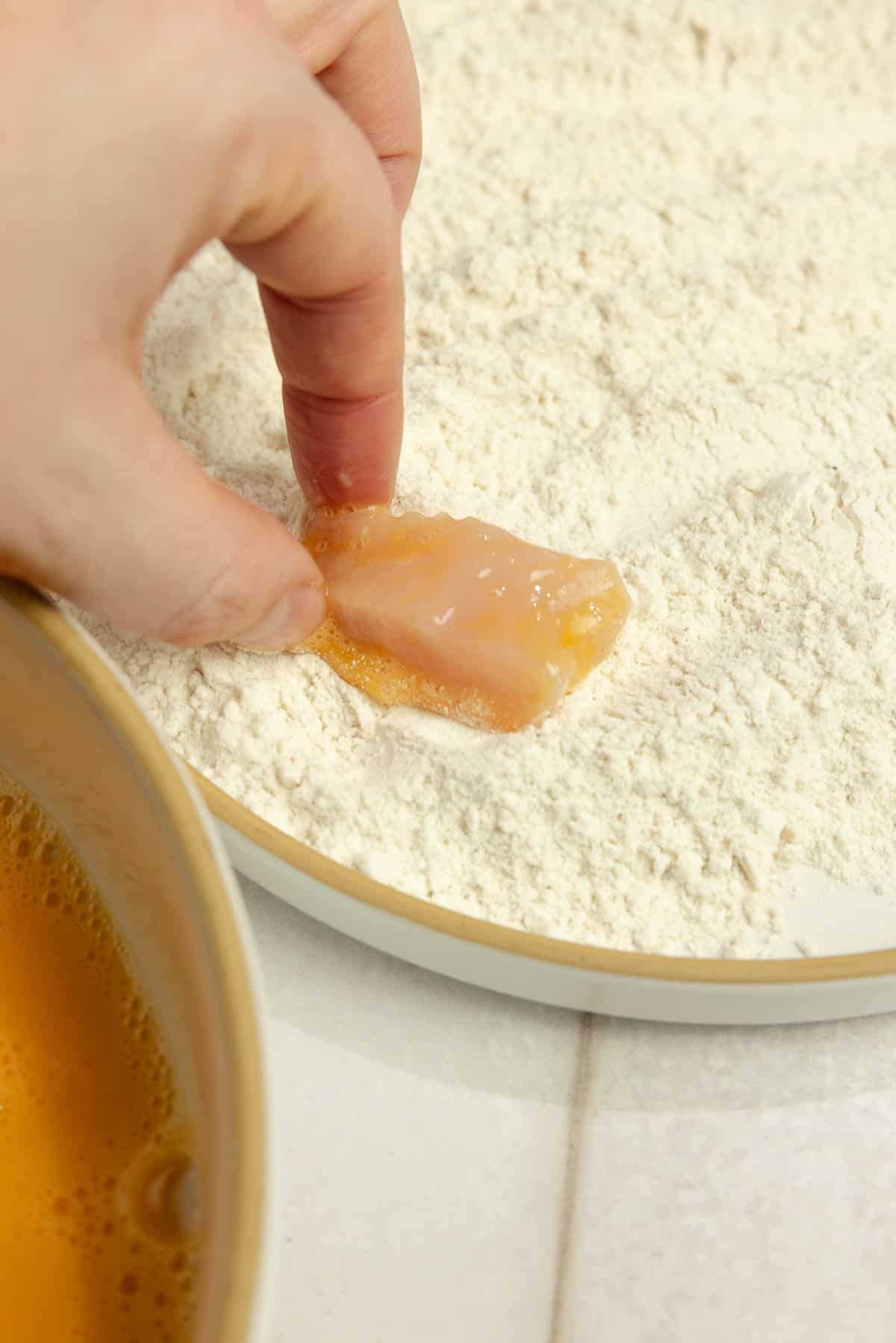 A hand dipping the egg-coated chicken into the flour.