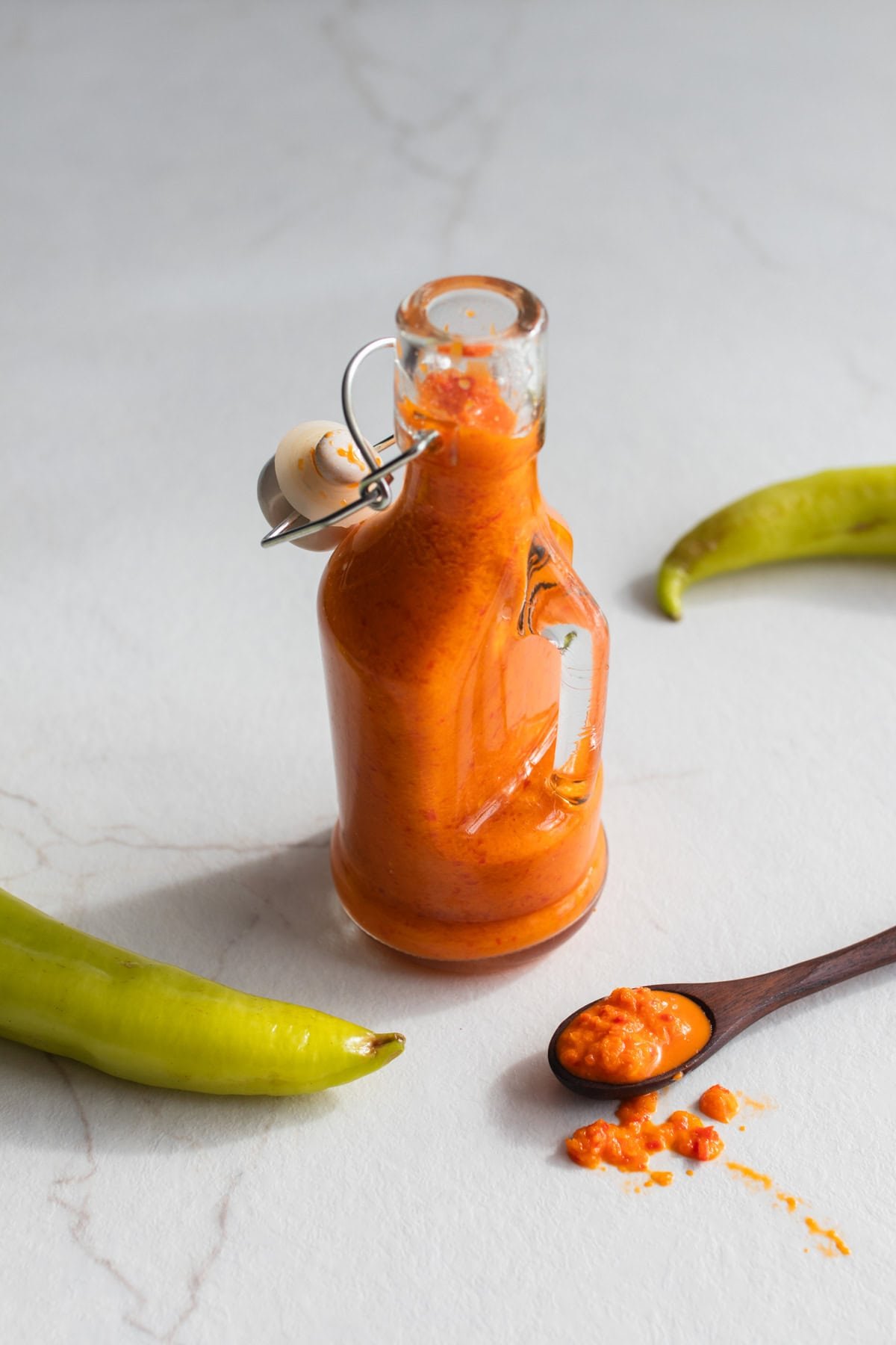 Fermented hot sauce in a glass bottle surrounded by hot peppers and a spoonful of hot sauce.