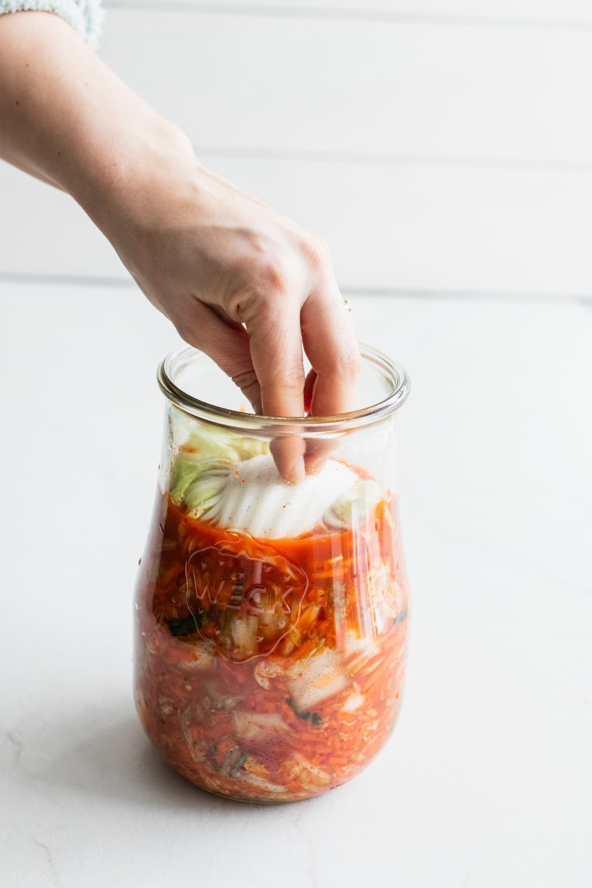 A hand folding cabbage leaves on top of kimchi in a jar.