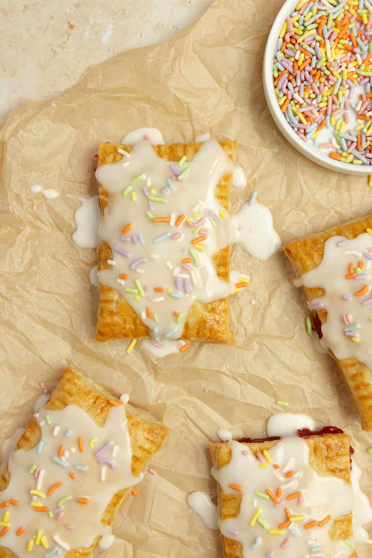 Four pop tarts topped with icing and sprinkles on parchment paper.