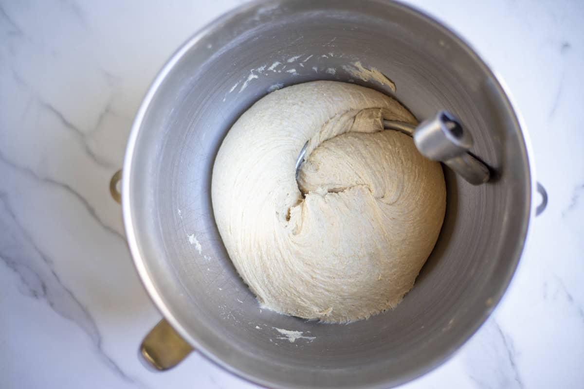 Bread dough in a bowl of a stand mixer with dough hook attachment.