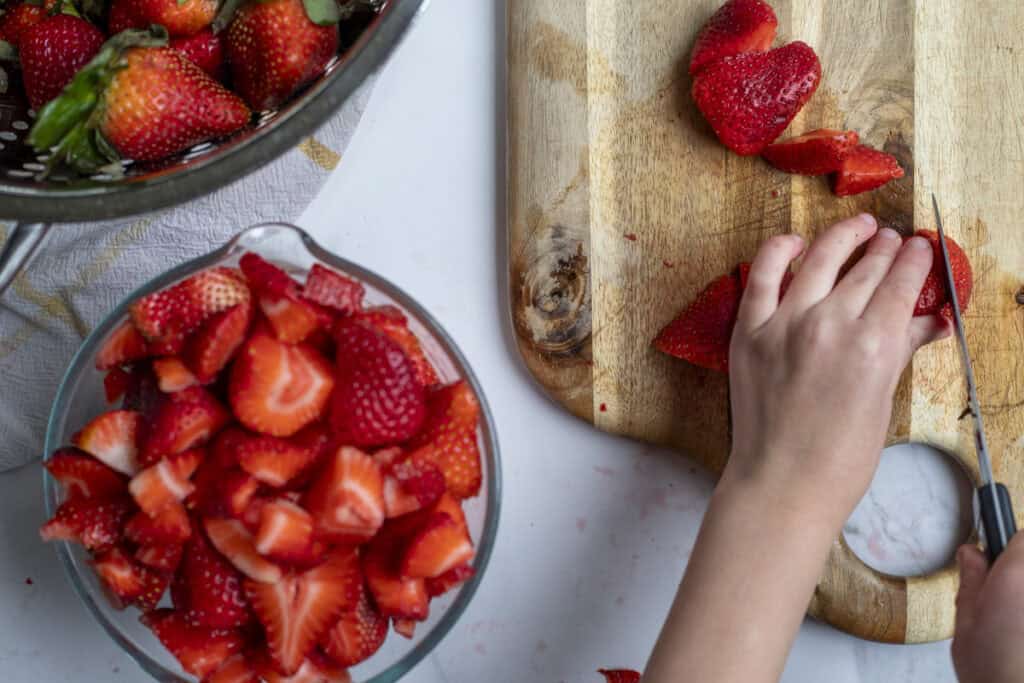 A hand cutting up strawberries on a cutting board.