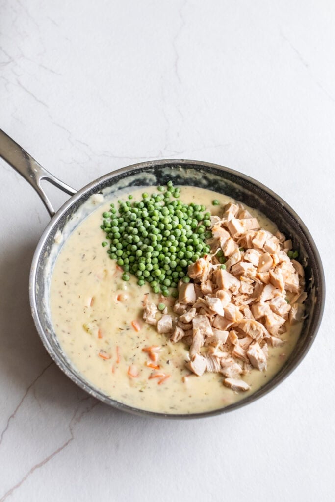 Chicken and peas being added to chicken pot pie filling in a skillet.