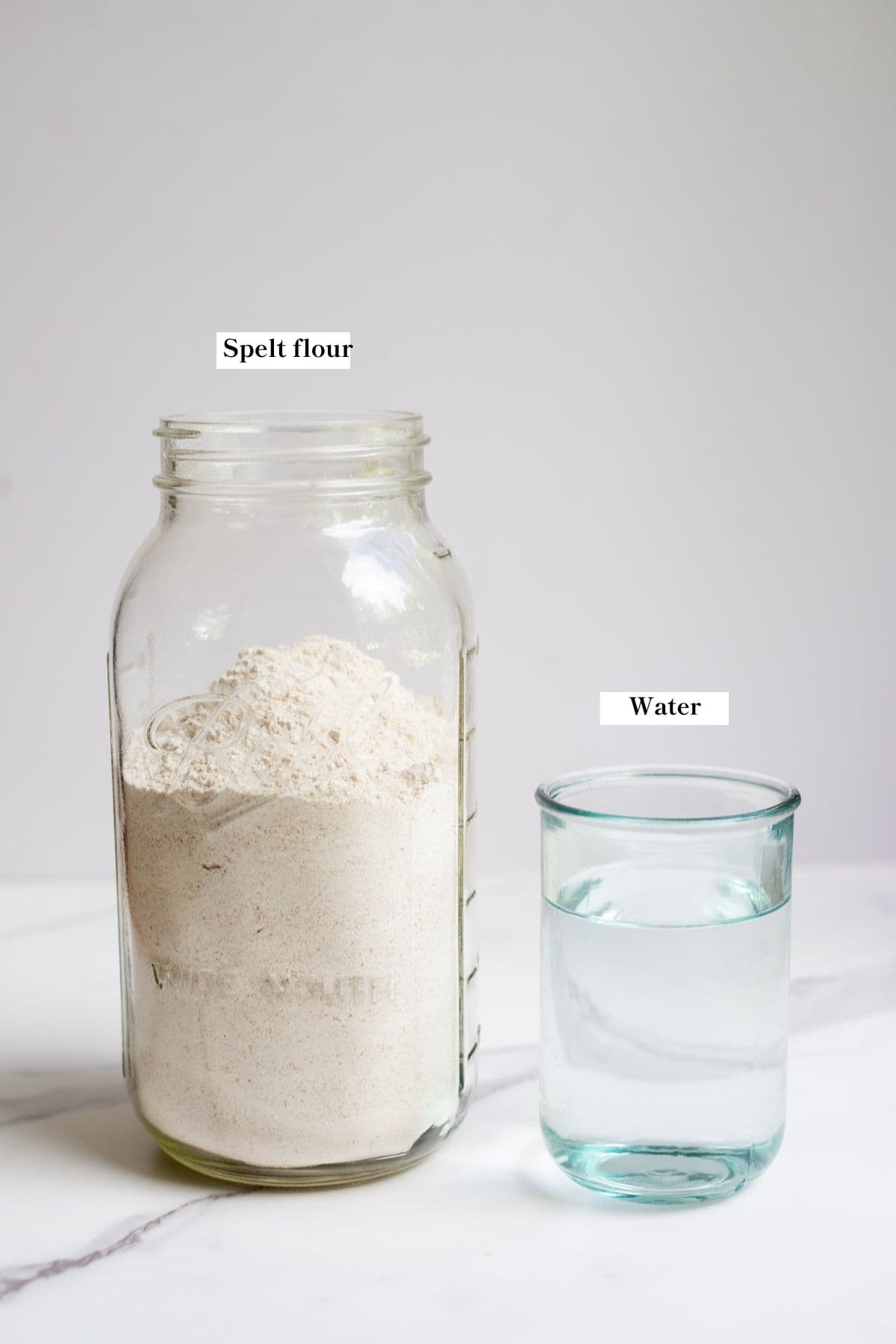 A half gallon mason jar of spelt flour and a glass of water on a white countertop.