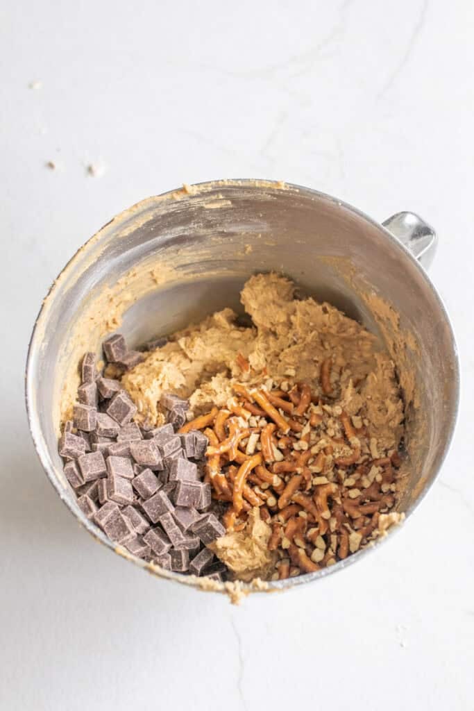Folding pretzels and chocolate chips into cookie dough in a bowl.
