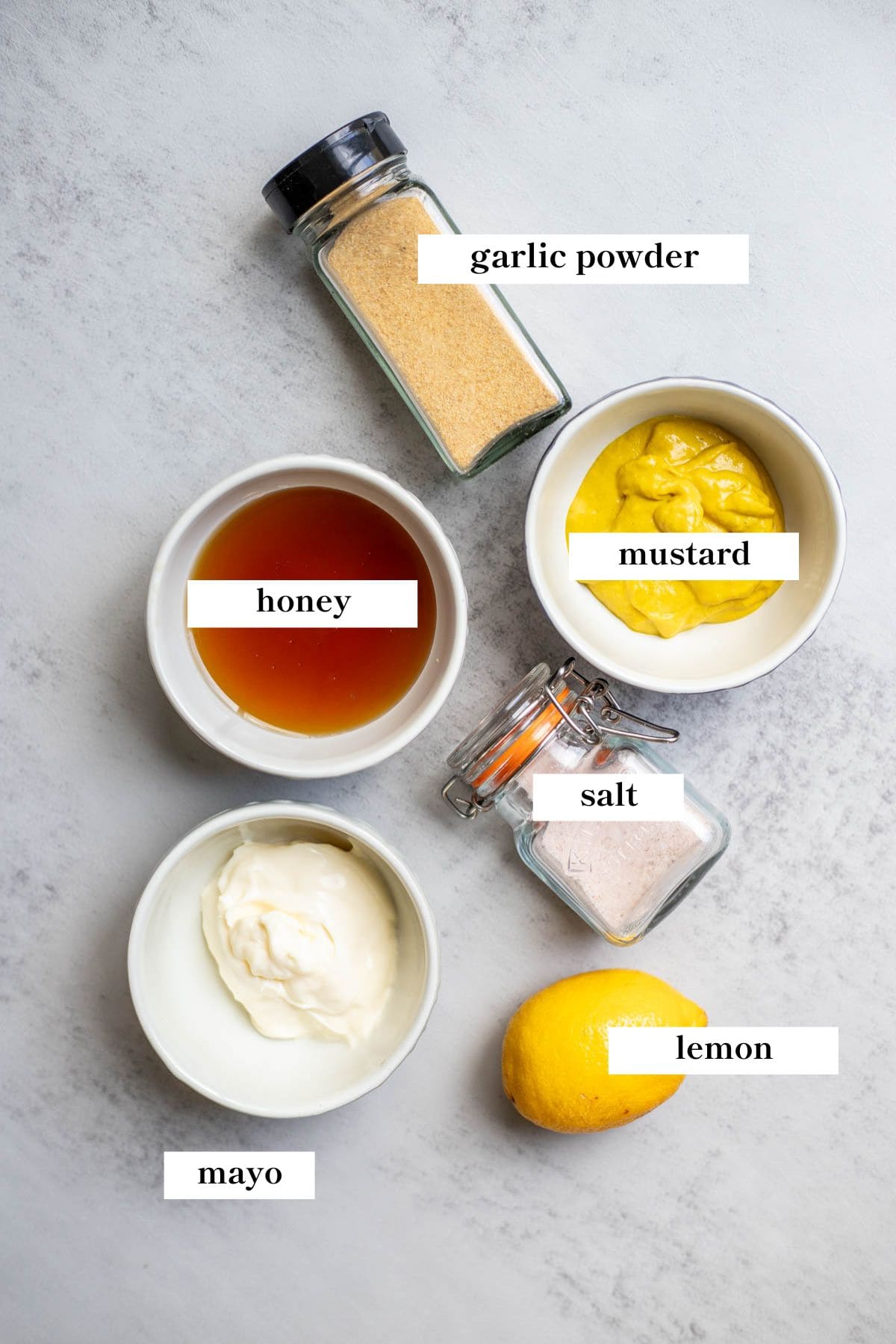 Small bowls of mayo, mustard, and honey, a lemon, salt, and a jar of garlic powder on a counter.