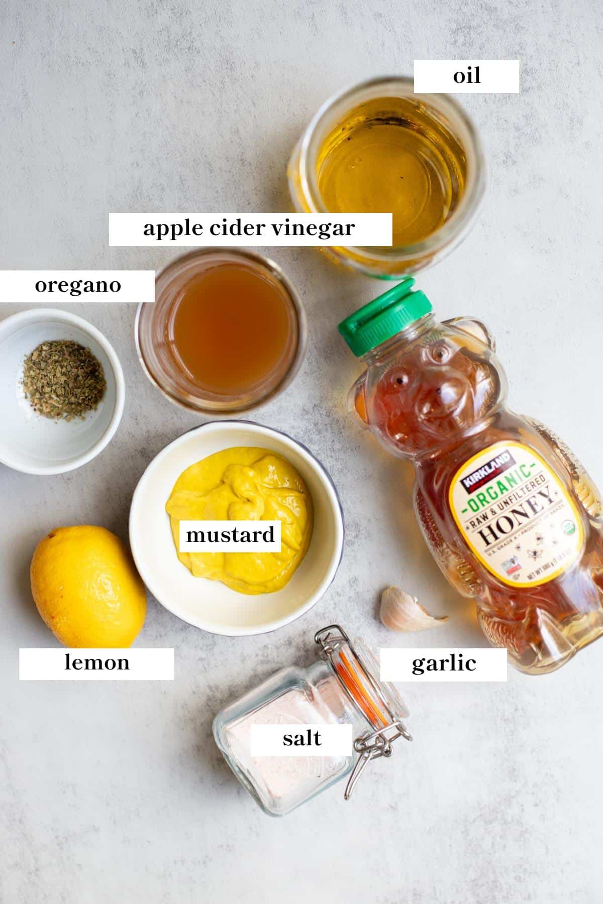 Oil, mustard, honey, salt, garlic, lemon, dried oregano and apple cider vinegar spread out on a countertop.