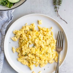 scrambled eggs on a white plate with a fork, fresh herbs and a salad are in the background.