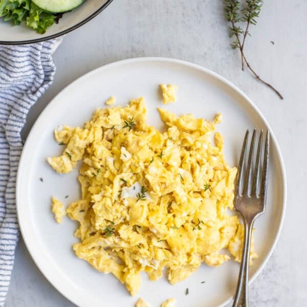 scrambled eggs on a white plate with a fork, fresh herbs and a salad are in the background.