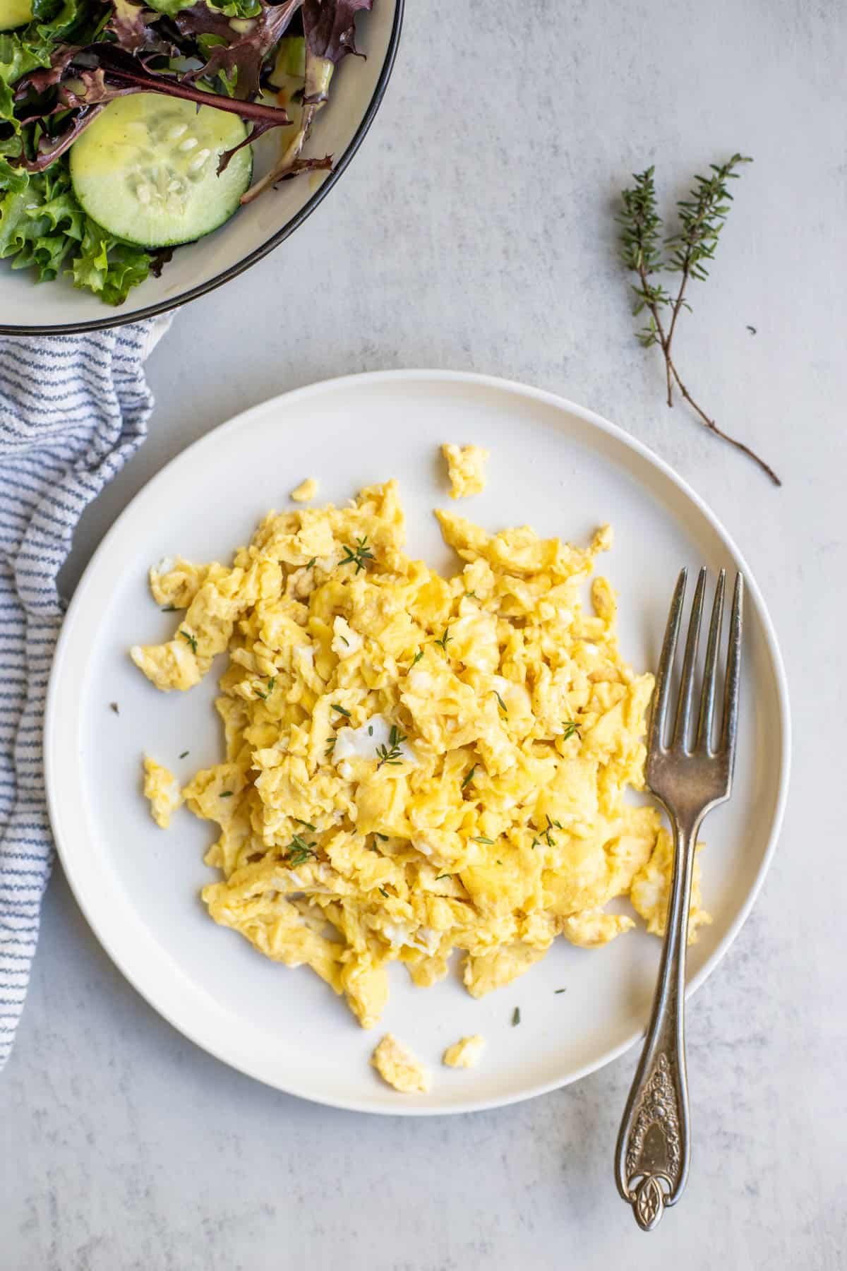 Scrambled eggs on a white plate with a fork, fresh herbs and a salad are in the background.