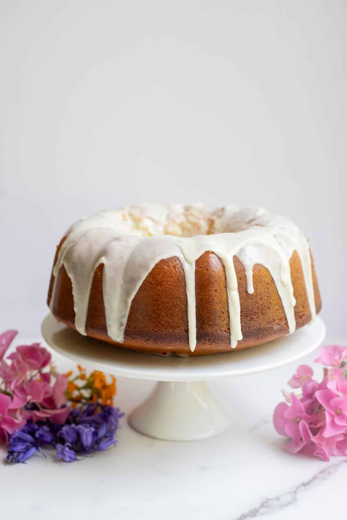 Sourdough Bundt cake with icing on a white cake stand surrounded by flowers.