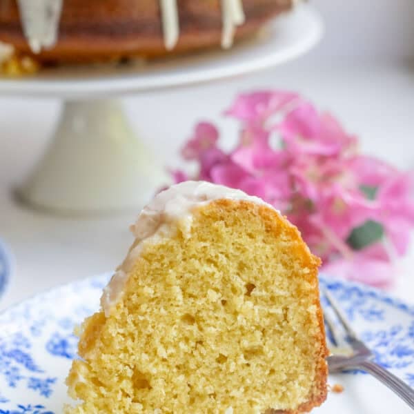 A slice of sourdough bundt cake topped with icing on a blue and white floral plate. The remaining cake is in the background on a cake pedestal.