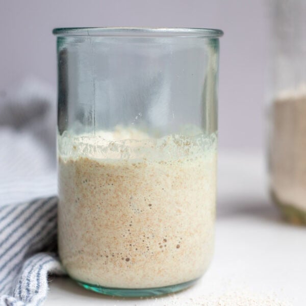 Spelt sourdough starter in a glass jar with a towel on a white countertop.