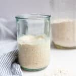 Spelt starter in a glass jar with a towel and another jar of flour.