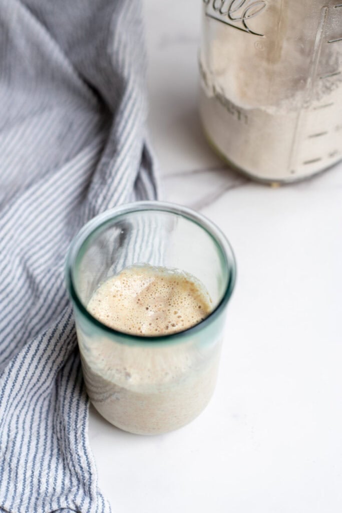 Spelt sourdough starter in a glass jar with a towel and a jar of spelt flour in the background.