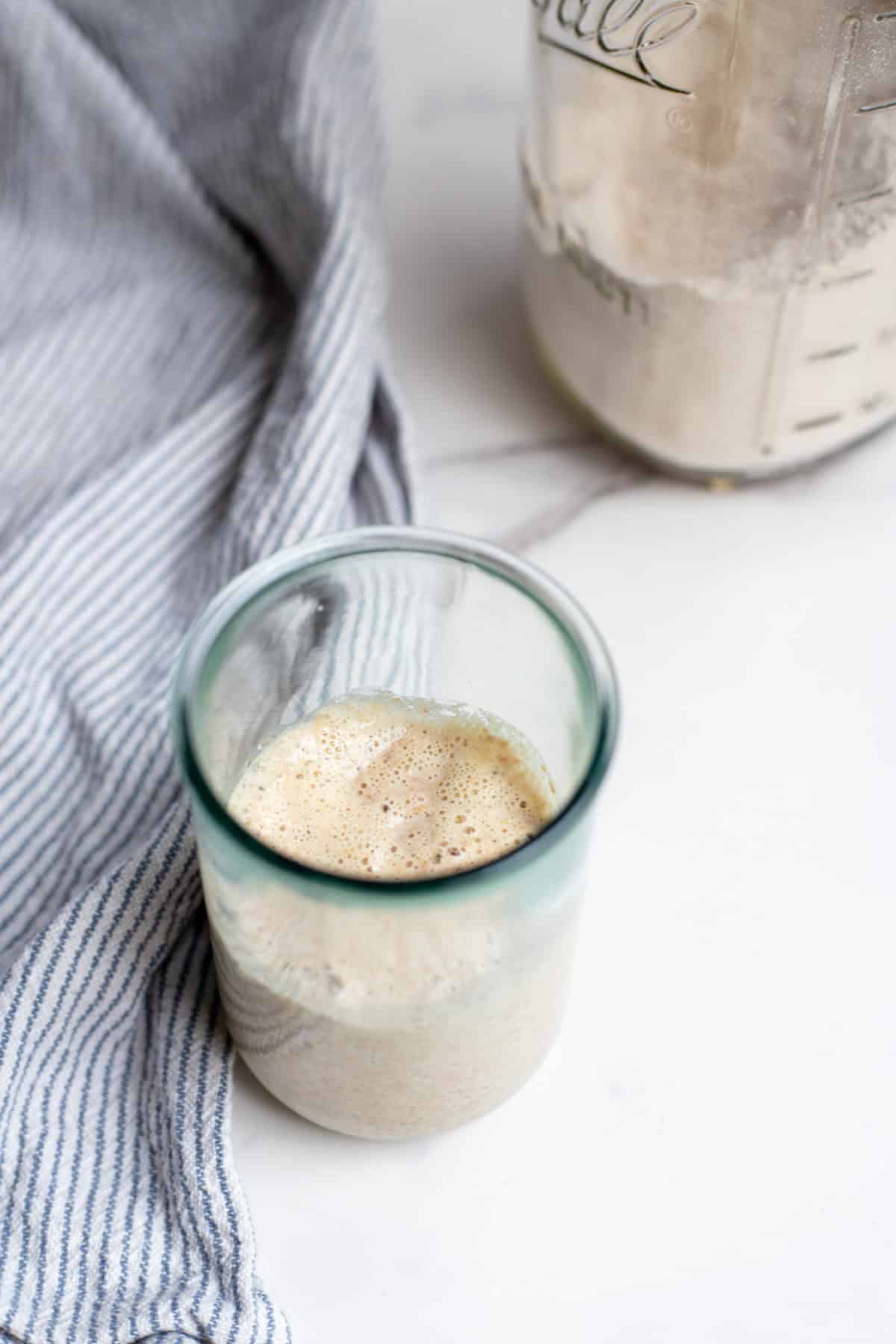Spelt sourdough starter in a glass jar with a towel and a jar of spelt flour in the background.